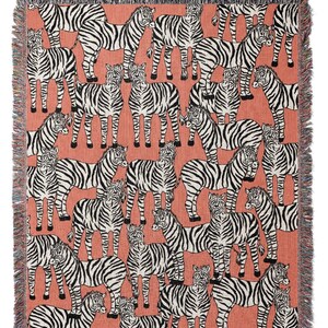 May include: Woven throw blanket featuring a repeating pattern of black and white zebras on a coral background. The blanket has a fringed edge in black, white, and coral. This decorative blanket is suitable for a bed or sofa.