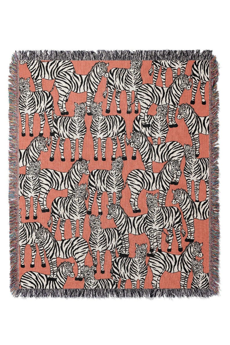 May include: Woven throw blanket featuring a repeating pattern of black and white zebras on a coral background. The blanket has a fringed edge in black, white, and coral. This decorative blanket is suitable for a bed or sofa.