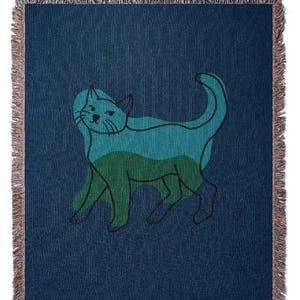 K&ouml;nnte beinhalten: Marineblaue Decke mit einem stilisierten Katzendesign. Die Katze ist in T&uuml;rkis- und Gr&uuml;nt&ouml;nen mit schwarzen Umrissen dargestellt. Die Decke hat einen Fransenrand.