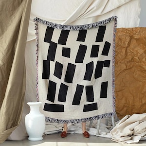 May include: A cream-colored throw blanket with a repeating pattern of black rectangular shapes. The blanket has a fringed edge and is displayed against a neutral backdrop. A white vase is in the foreground.