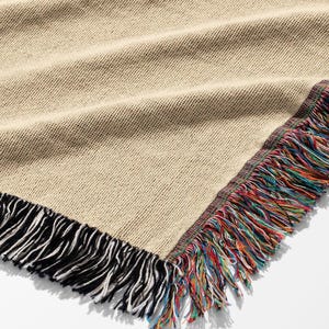 K&ouml;nnte beinhalten: Beige gewebte Decke mit farbenfrohem Fransenrand. Die Decke hat ein strukturiertes Aussehen und einen schwarz-wei&szlig;en Fransen an einer Seite. Die andere Seite hat einen lebendigen, mehrfarbigen Fransen.