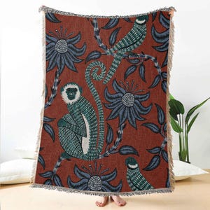 Op de afbeelding: Geweven plaid met een herhalend patroon van apen, vogels en bloemen in teal en grijs op een roestkleurige achtergrond. De deken heeft een gefranjerde rand en is gemaakt van een zacht, getextureerd materiaal. Het ontwerp doet denken aan een vintage botanische illustratie.