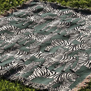 May include: A green throw blanket with a repeating pattern of black and white zebras. The blanket has a fringed edge and is displayed on a bed of green grass. The zebras are in various poses.
