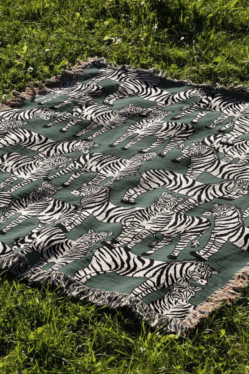 May include: A green throw blanket with a repeating pattern of black and white zebras. The blanket has a fringed edge and is displayed on a bed of green grass. The zebras are in various poses.