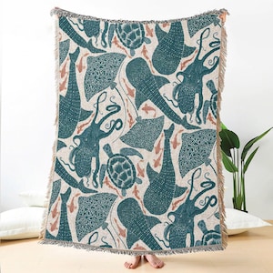 May include: A woven throw blanket with a teal marine life design on a cream backdrop. The pattern showcases octopuses, turtles, stingrays, whale sharks, and fish. The blanket has a fringed edge and is made of a soft, woven material.