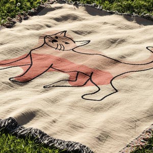 K&ouml;nnte beinhalten: Beige Decke mit einer gro&szlig;en Katzenillustration in Schwarz und Koralle. Die Katze liegt mit eingerolltem Schwanz. Die Decke hat einen Fransenrand und liegt auf gr&uuml;nem Gras.