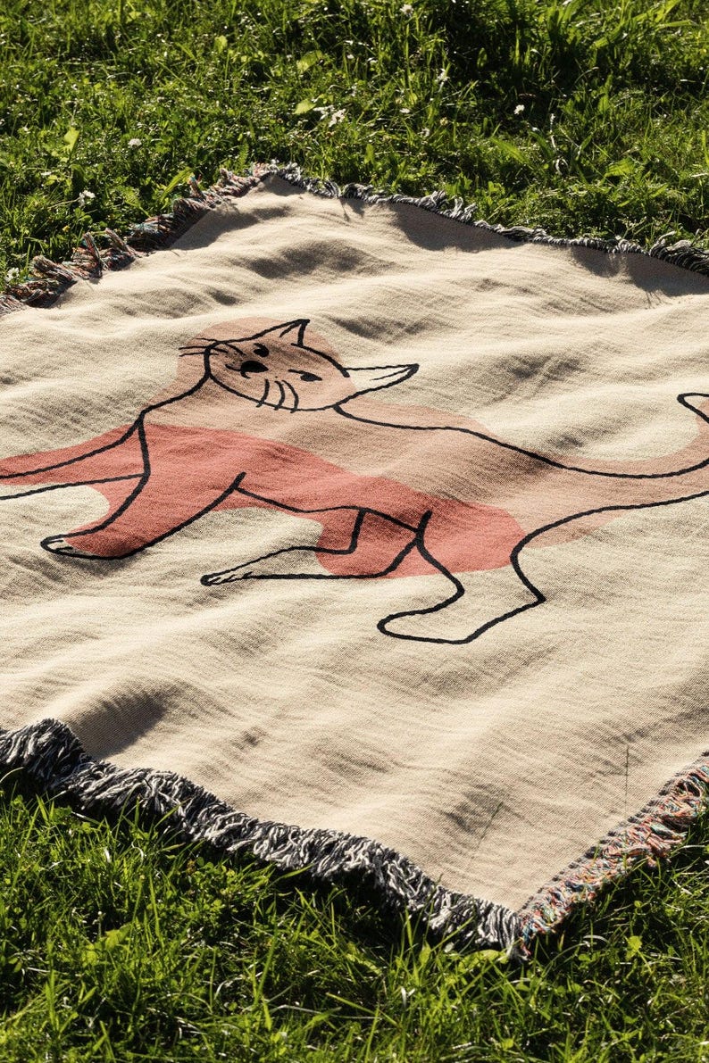 K&ouml;nnte beinhalten: Beige Decke mit einer gro&szlig;en Katzenillustration in Schwarz und Koralle. Die Katze liegt mit eingerolltem Schwanz. Die Decke hat einen Fransenrand und liegt auf gr&uuml;nem Gras.