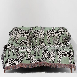 May include: A green throw blanket featuring a repeating pattern of black and white zebras. The blanket is draped over a couch and has a fringed edge with a mix of colors. The design is a playful take on animal print.
