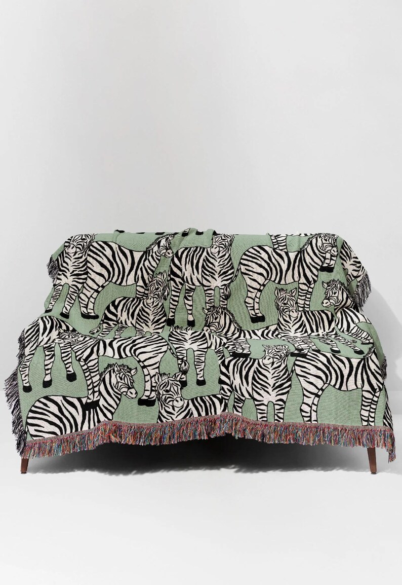 May include: A green throw blanket featuring a repeating pattern of black and white zebras. The blanket is draped over a couch and has a fringed edge with a mix of colors. The design is a playful take on animal print.