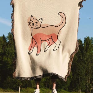 K&ouml;nnte beinhalten: Cremefarbene Wurkdecke mit einer Katzenillustration. Die Katze ist schwarz umrandet mit Pfirsich- und hellbraunen Akzenten. Die Decke hat einen Fransenrand und wird vor blauem Himmel und gr&uuml;nen B&auml;umen gehalten.