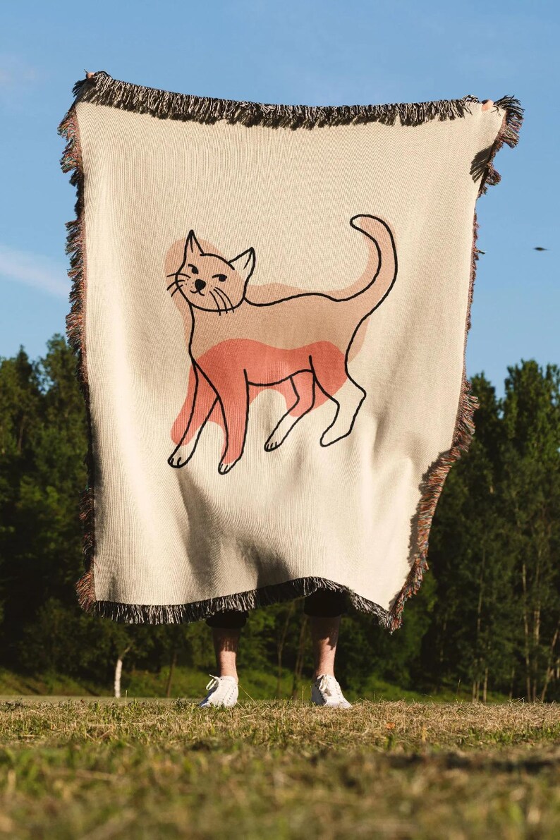K&ouml;nnte beinhalten: Cremefarbene Wurkdecke mit einer Katzenillustration. Die Katze ist schwarz umrandet mit Pfirsich- und hellbraunen Akzenten. Die Decke hat einen Fransenrand und wird vor blauem Himmel und gr&uuml;nen B&auml;umen gehalten.