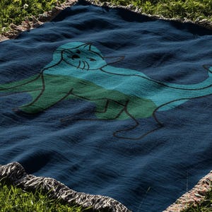 K&ouml;nnte beinhalten: Eine marineblaue Decke mit einer Katzenillustration in Blau- und Gr&uuml;nt&ouml;nen. Die Decke hat einen Fransenrand und liegt auf gr&uuml;nem Gras.