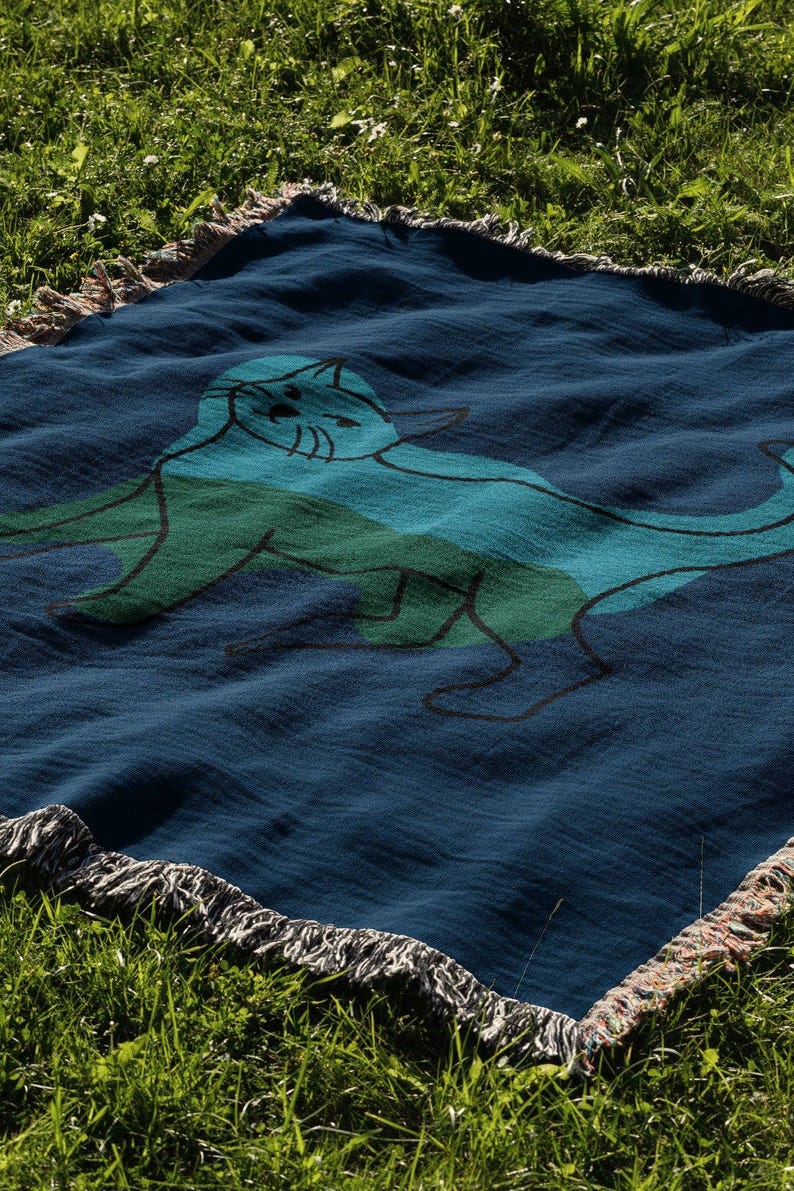 K&ouml;nnte beinhalten: Eine marineblaue Decke mit einer Katzenillustration in Blau- und Gr&uuml;nt&ouml;nen. Die Decke hat einen Fransenrand und liegt auf gr&uuml;nem Gras.