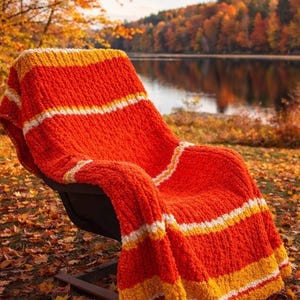 Puede incluir: Una manta de punto a rayas naranja y blanca, sobre una silla negra. La manta es gruesa y texturizada, con bandas blancas y amarillas. La escena es al aire libre, con hojas de otoño y un lago al fondo.