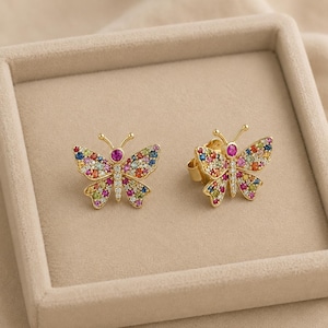 May include: A pair of gold butterfly stud earrings featuring wings encrusted with multicolored gemstones and a central body accented with clear white stones. Each butterfly has a prominent pink stone at the head and delicate antennae.