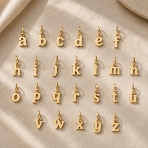 May include: A collection of twenty-six individual gold-toned metal charms, each shaped as a lowercase letter of the alphabet from a to z. Every charm features a small circular jump ring at the top for attachment, arranged in four rows.