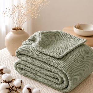 Pode incluir: Uma manta verde sálvia com um padrão de tecido waffle texturizado, dobrada cuidadosamente numa pilha. O tecido apresenta uma estrutura de grelha consistente em toda a superfície, exibindo um tom verde suave e um aspeto espesso e em camadas.