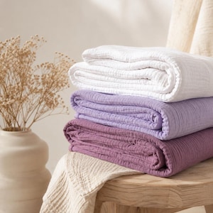 Pode incluir: Uma pilha de três mantas de musselina dobradas nas cores branco, lavanda claro e ameixa escuro. As mantas apresentam uma superfície de tecido texturizado e enrugado. Uma manta adicional cor de creme está pendurada num banco de madeira.