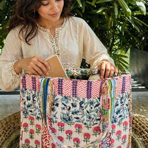 Könnte beinhalten: Eine große, farbenfrohe Tragetasche mit Patchwork-Design. Die Tasche zeigt Blumen- und geometrische Muster in Rosa, Blau und Weiß. Sie hat stabile Griffe und ist oben offen, wodurch das Innere sichtbar wird. Die Tasche steht auf einer Korboberfläche.
