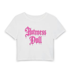 以下が含まれることがあります： ピンク色の「Hotmess Doll」の文字が滴るフォントでプリントされた白いクロップド丈トップス。クルーネックと半袖のデザインです。テキストはシャツの前面中央に配置されています。
