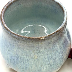 May include: A handmade ceramic bowl with a light blue and white glaze. The bowl has a rounded shape with a slightly flared rim and a brown rim. The exterior has a textured, dripped appearance.