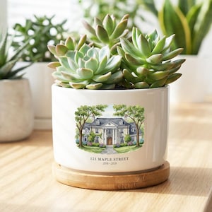 May include: White ceramic planter with a watercolor illustration of a house, trees, and the text "123 Maple Street 2016-2020". The planter holds green succulents and sits on a wooden coaster. Other potted plants are in the background.