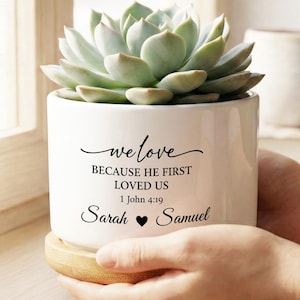 May include: A white ceramic planter with a succulent plant, held by hands. The planter has the text "we love because he first loved us" and the names Sarah and Samuel. The planter sits on a wooden coaster.