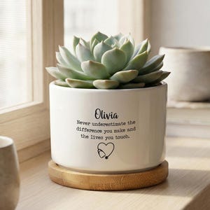 May include: A white ceramic plant pot with a succulent, on a wooden coaster. The pot is inscribed with the name "Olivia", a quote about making a difference, and a stethoscope heart symbol.