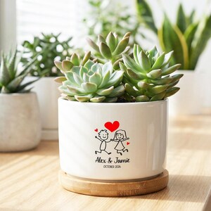 May include: A white ceramic planter with a succulent plant, featuring a heart and stick figure couple design. The planter includes the names "Alex & Jamie" and the date "October 2026". The planter sits on a wooden coaster, with other plants in the background.