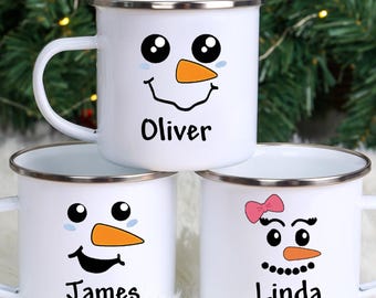 May include: Three white enamel mugs with snowman faces and names. Each mug has a black smiling face, black eyes, and an orange carrot nose. The names "Oliver", "James", and "Linda" are printed below the faces.