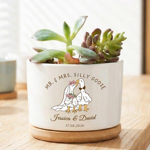 以下が含まれることがあります： ウェディングドレスを着た2羽のガチョウのイラストが描かれた、竹の受け皿付きの白いセラミック植木鉢。多肉植物が入っており、「Mr. & Mrs. Silly Goose」の文字と結婚式の日付が書かれています。