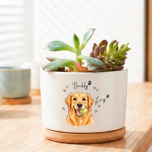 May include: A white ceramic planter with a watercolor illustration of a Golden Retriever holding a tennis ball. The planter has the names "Buddy" and "Lucy" and is filled with succulents. It sits on a wooden coaster.