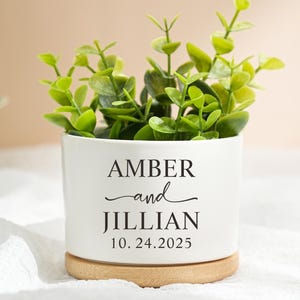 May include: A white ceramic planter with the names "AMBER and JILLIAN" and the date "10.24.2025" in black lettering. The planter holds green leafy plants and sits on a round wooden base.