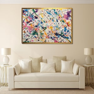 May include: Abstract art painting with a gold frame. The artwork features a white background with colorful paint splatters in shades of blue, pink, purple, green, and yellow. The painting is displayed above a cream-colored sofa with pillows and two white lamps.