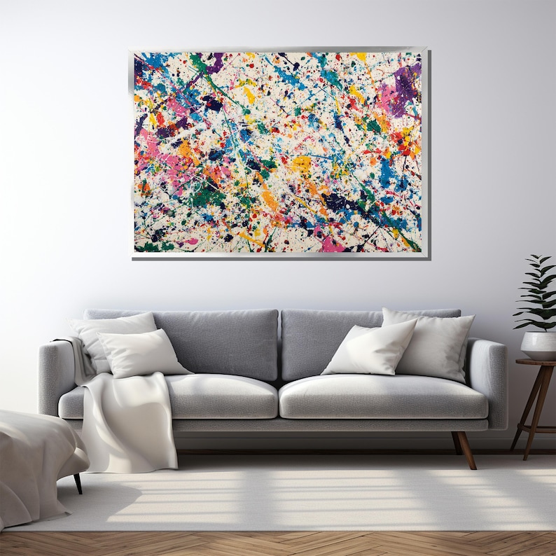 May include: Abstract art piece with a white background and colorful paint splatters in shades of blue, pink, yellow, green, and purple. The artwork is framed and mounted on a white wall above a gray sofa.