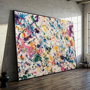 Könnte beinhalten: Großformatiges abstraktes Gemälde mit schwarzem Rahmen. Das Kunstwerk zeigt einen weißen Hintergrund mit bunten Farbspritzern in Blau, Grün, Gelb, Orange, Rosa und Lila. Das Gemälde lehnt an einer grauen Wand.