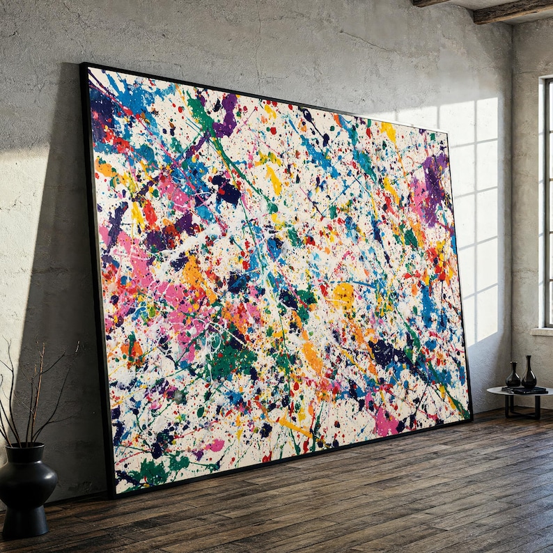 May include: Large abstract painting with a black frame. The artwork features a white background covered in colorful paint splatters of blue, green, yellow, orange, pink, and purple. The painting is leaning against a gray wall.