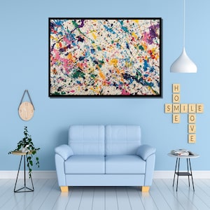 May include: Abstract art print with a black frame. The artwork features a white background covered in colorful paint splatters of blue, pink, yellow, green, and purple. The painting hangs above a light blue sofa.