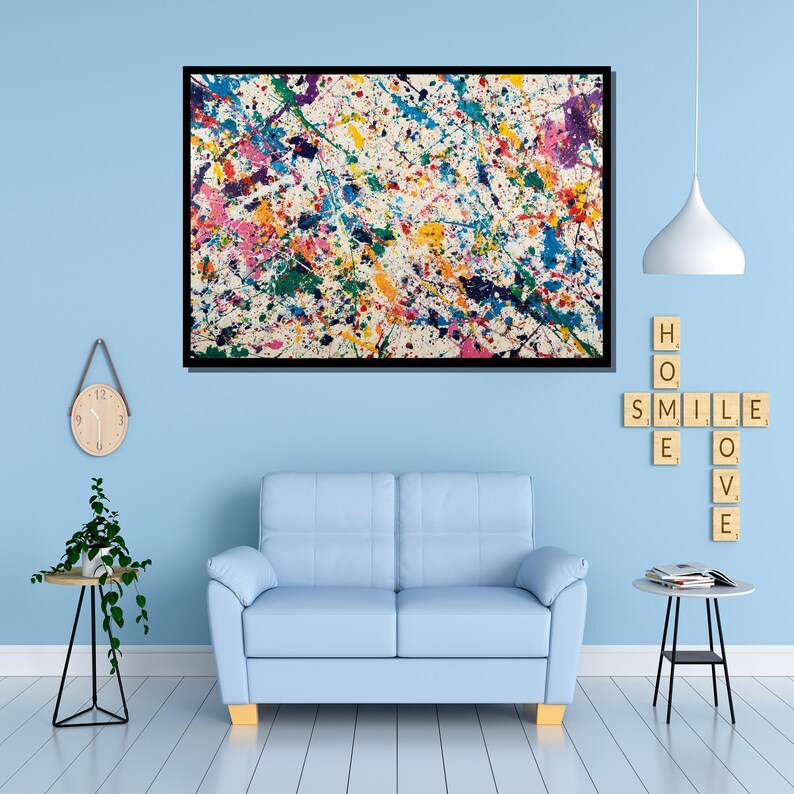 May include: Abstract art print with a black frame. The artwork features a white background covered in colorful paint splatters of blue, pink, yellow, green, and purple. The painting hangs above a light blue sofa.