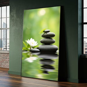 May include: A framed print featuring a zen-inspired scene. A stack of smooth, dark gray stones is balanced atop a reflective surface, with a white lotus flower and green foliage. The background is a soft, blurred green.