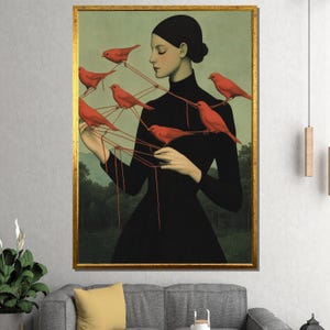 May include: A framed art print featuring a woman in a black dress with red birds connected by strings. The artwork has a gold frame and a muted green and black background. The birds are a vibrant red color.