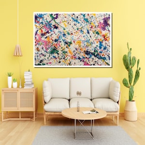 May include: Abstract art piece with a white background and colorful paint splatters in shades of blue, pink, yellow, green, and purple. The artwork is framed and hangs above a white sofa and wooden coffee table.