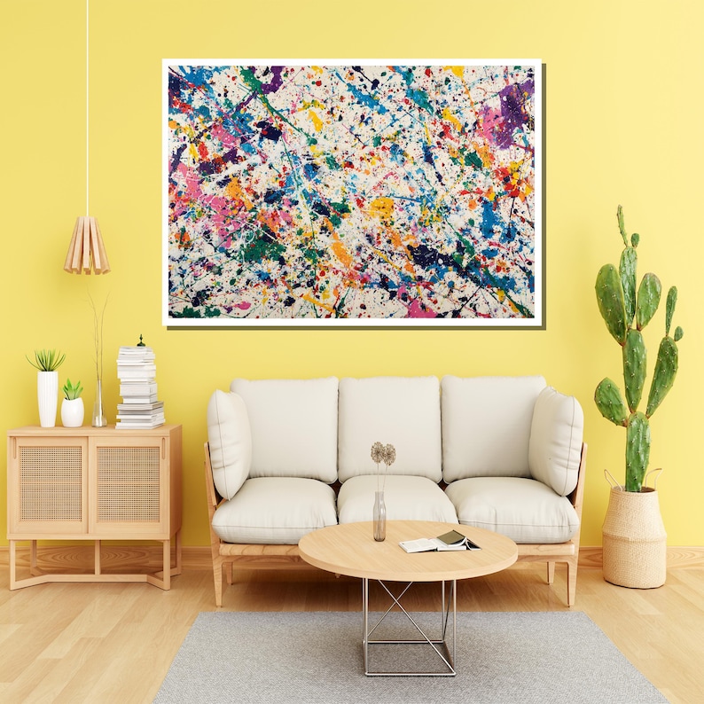 May include: Abstract art piece with a white background and colorful paint splatters in shades of blue, pink, yellow, green, and purple. The artwork is framed and hangs above a white sofa and wooden coffee table.