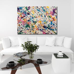 May include: Abstract art canvas with a white background and colorful paint splatters in shades of blue, green, pink, yellow, and purple. The artwork is displayed above a white sofa and glass coffee table.