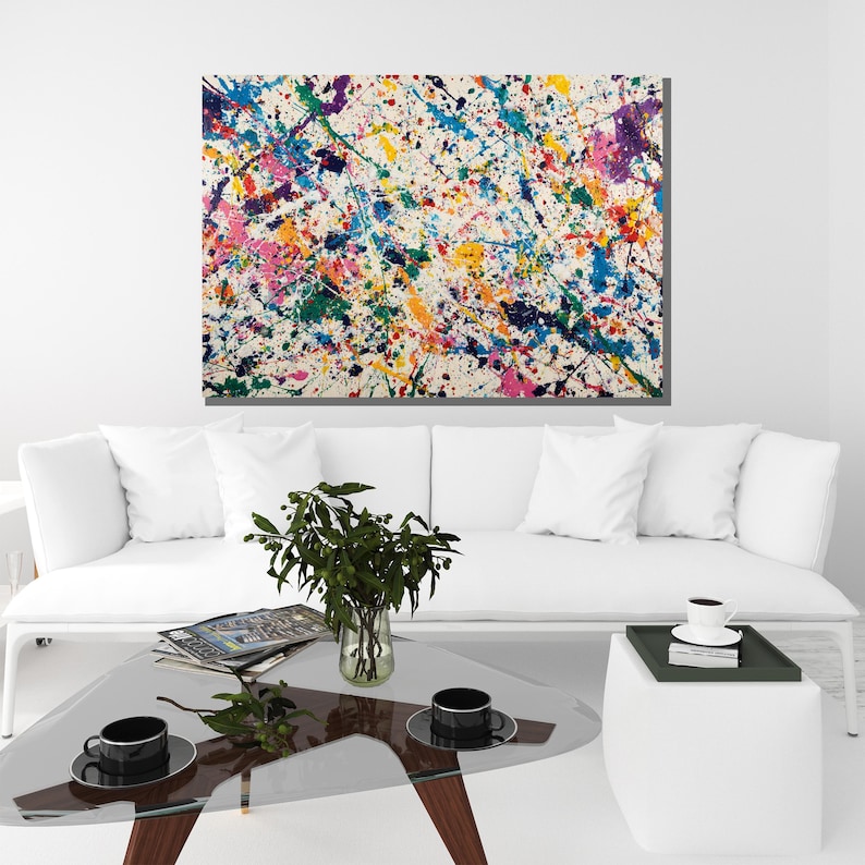 May include: Abstract art canvas with a white background and colorful paint splatters in shades of blue, green, pink, yellow, and purple. The artwork is displayed above a white sofa and glass coffee table.