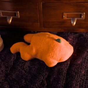May include: An orange, abstract-shaped object with a small green accent, possibly a decorative item. The object rests on a dark, textured surface, with a wooden cabinet and a white, spiral candle in the background.