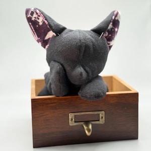 May include: A black plush cat toy with patterned purple and white ears, resting in a small wooden box with a brass label holder. The cat is posed with its paws resting on the box.