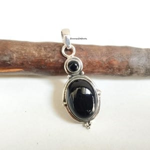 May include: A silver pendant with a large, oval black stone and a smaller, round black stone above it. The pendant is set against a wooden background. The pendant has a loop at the top for a chain.