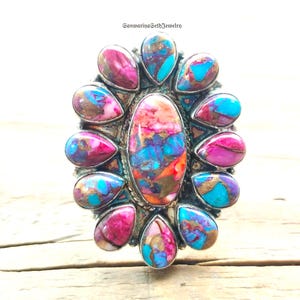 Puede incluir: Un anillo ovalado con un conjunto de piedras de colores. El anillo presenta una piedra central grande rodeada de piedras más pequeñas en forma de lágrima en tonos rosa, turquesa y dorado. El anillo está engarzado en una banda plateada.