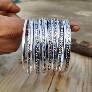 Könnte beinhalten: Eine Sammlung silberner Armreifen mit aufwendigen floralen und geometrischen Mustern. Die Armreifen werden in einer Hand präsentiert, die die detaillierten Designs und die polierte Oberfläche zeigt. Die Armreifen bestehen aus glänzendem, silbernem Metall.